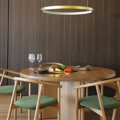 Contemporary lightwood, dining table that is round in shape with contemporary wooden chairs and crescent bench, and decorated with wine glasses, cutlery and a simple cheeseboard that is lit up by a decorative low-lying, golden 'halo' light. Ebb Tide.