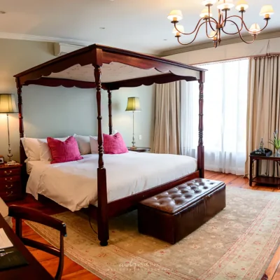 Bedroom suite in Cape Heritage Hotel with an antique wooden four poster bed with crisp white linen and pink cushions and complimentary wooden side tables set against an egg blue washed wall that reflects in the Persian rug.