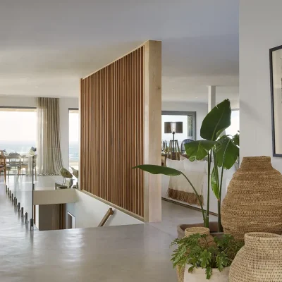 An upper level dining and living area in Walkers Beach Villa with grey epoxy flooring, a wooden cladded statement wall and a wooden dining table facing the expansive glass doors that open the space to views of the blue ocean.