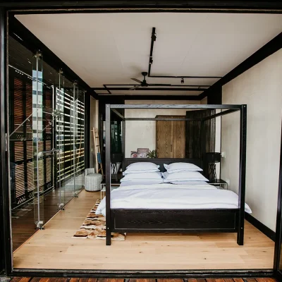 A wooden four poster bed with white crisp linen , two wooden side tables and a soft fur rug on hard wood flooring in The View bedroom suite surrounded by large glass doors that open onto an enclosed wooden deck.