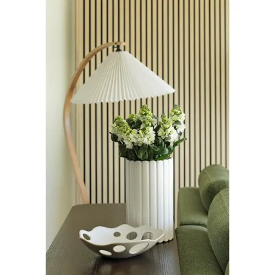 A tall, white vase containing fresh, white flowers that sits on a wooden cabinet in front of Ebb Tide's contemporary, striped wall paper with a freestanding, curved lamp.