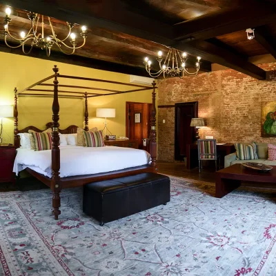 Bedroom suite with a wooden four poster bed and crisp white linen positioned against a yellow statement wall, a large Persian rug exposing 200 year old Cape yellowwood floors, and a soft beige sofa positioned against the original plasterwork. Cape Heritage Hotel.