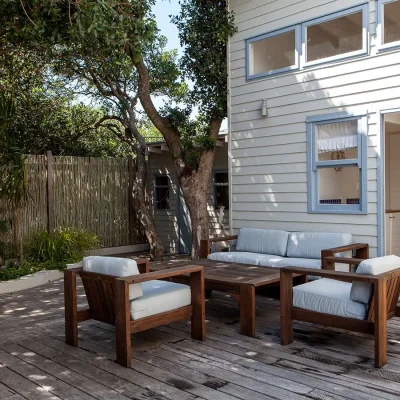 Secluded back yard deck with wooden outdoor furniture and luscious trees providing shade. Pebbles.