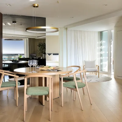 Open plan dining area in Ebb Tide beach abode, that is furnished with a sleek, rounded light wooden dining table and chairs, and sits in a light exposing corner of a mirrored wall and stack-back wooden doors.