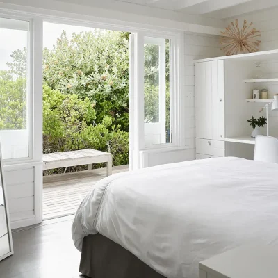 Noordhoek Beach House bedroom with white built in cupboards and desk, and a spacious crisp white linen bed facing glass double doors that open onto a wooden deck with views of the indigenous garden.