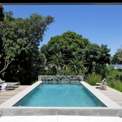 Blue lap pool surrounded by a wooden deck with sun loungers and protected by the luscious green garden that exposes gaps of Noordhoek beach. Pinebeach.