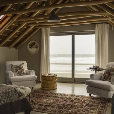 Main bedroom suite with thatched roofing in Beach house Zonnestraal furnished elegantly with a comfortable bed facing two large soft, white armchairs and glass sliding doors that expose views of the ocean.