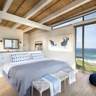 Bedroom suite with a beige floating wall separating the large bed with crisp white linen and blue textiles from the bathroom with a large shower that looks out onto views of the ocean from the large glass windows. Lekkerwater Beach Lodge.