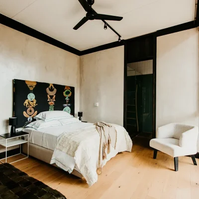 Bedroom suite with beige washed walls and hard wood floors, a spacious bed with luxury white linen and beige throw set against a black headboard with decorative illustrations, a corner arm chair and an ensuite bathroom.