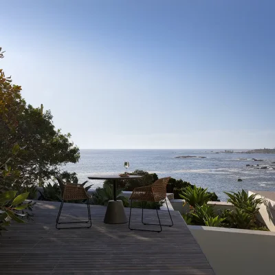 Elongated wooden deck with a table and two chairs that looks onto the shimmering ocean and shaded by the luscious green trees.