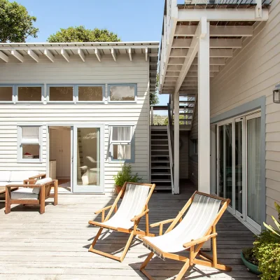 Beige clapboard home with double doors that open onto Pebbles wrap around deck with wooden sofas and sun loungers.