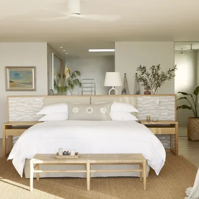 Open plan bedroom suite with ensuite bathroom, and furnished with a luxurious white linen bed, modern wooden side tables, a wicker ottoman at the foot of the bed and a large mirror. Walkers Beach Villa.