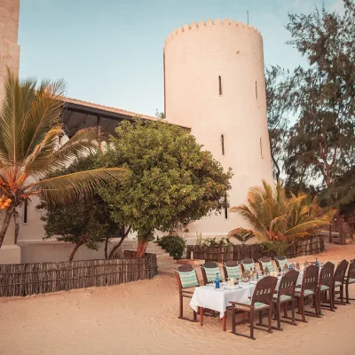 Extensive wooden dining table and chairs set with white linen cloth that matches the white beach sand and the Fort in the background.