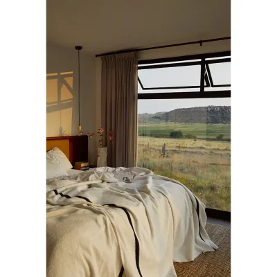 Bedroom with a mess of soft linen on the bed and warmed by the golden morning sunlight seeping through the large glass windows to expose views of the vast grassy plains. The Herenberg.