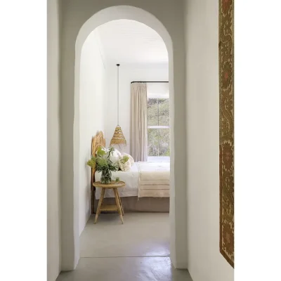 Arched entrance with whitewashed walls revealing a spacious bed with white linen, a wicker headboard and side table. Buchubos Villa.