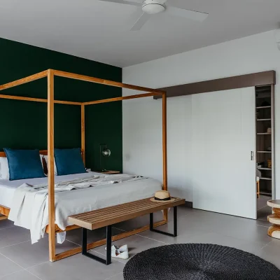 Modern but simple room in Black river Penthouse with large wooden four poster bed with white linen against an emerald green wall, two bedside tables, a wooden ottoman at the foot of the bed, and a soft, beige armchair.