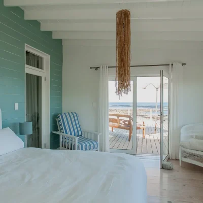 Bedroom in Pebbles home with a turquoise statement wall, a spacious white linen bed positioned below a decorative hanging light and double glass doors that open onto a wooden deck with views of the ocean.