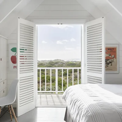 Loft bedroom with white cladded walls and ceiling, built in cupboards and a desk, a spacious white linen bed, and double doors that open onto a wooden deck with views of the coastal vegetation and ocean.