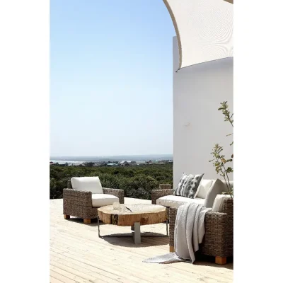 Outdoor lounging on a sweeping deck with woven armchairs and white pillows surrounding a statement coffee table made of solid wood, and facing coastal vegetation and the crystal blue ocean.