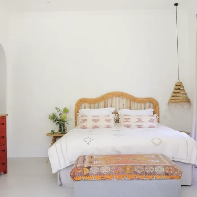 Buchubos Villa bedroom with whitewashed walls, a spacious bed with white linen and touches of pink attached to a wicker headboard, a side table and a colourful fabric ottoman at the head of the bed, with a red statement cabinet.