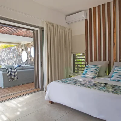 Comfortable bed set against a wooden panelled wall with an air conditioner, and opens onto outdoor deck via large sliding doors.