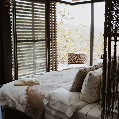 Luxurious bedroom with white linen, and decorative pillows and a beige throw overlooking large stack back glass doors with a corner leopard print armchair.