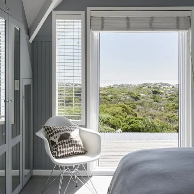 Glass double doors exposing views of the coastal vegetation and ocean from a Noordhoek Beach House bedroom with soft grey cladded walls and built in cupboards with mirrors attached.