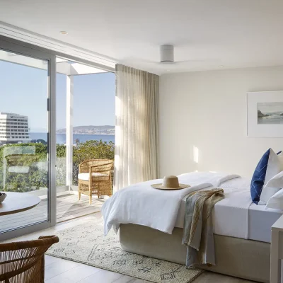 Comfortable bedroom suite with luxurious white linen bed, deep blue scatter pillows, white wooden side tables, a corner table with wicker armchairs, and large glass sliding doors that open onto a sea facing deck. Walkers Beach Villa.