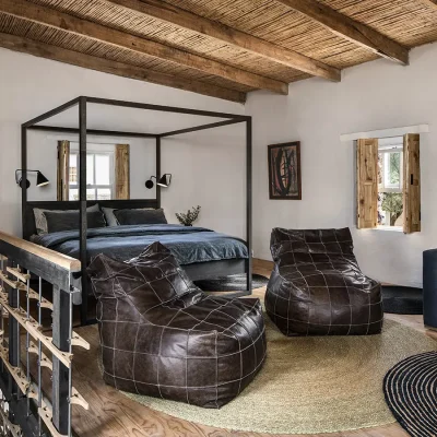 Open-plan room with large four poster bed with navy and charcoal linen faces two leather ottomans.