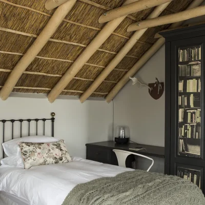 Cosy single bed with white linen and floral pillows, a side table and a large wooden cupboard with decorative black and white wallpaper, and a tucked away corner desk. Beach House Zonnestraal.