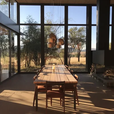 Extensive wooden dining table and chairs positioned below silver bulb hanging lights and adjacent to a large indoor fireplace that is enclosed by large glass walls that expose the outdoor bushveld. Witklipfontein ecolodge.