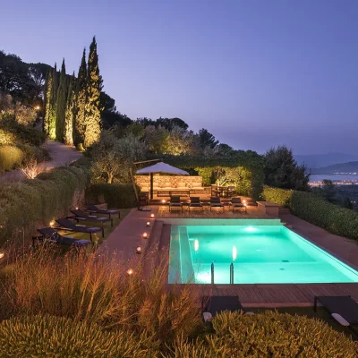 Evening at Villa Sunshine with a lit up swimming pool area, and a surrounding garden lit up by foot path lights, and a purple sky with distant ocean views.