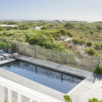 Swimming pool surrounded by a wooden deck and coastal vegetation. Noordhoek Beach House.