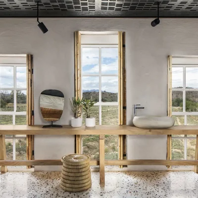 Long and narrow wooden bench with stone washbasin stretches across three large windows with wooden shutters overlooking Karoo vegetation and mountains, and has granite flooring with a cream finish. Buffelsdrift.