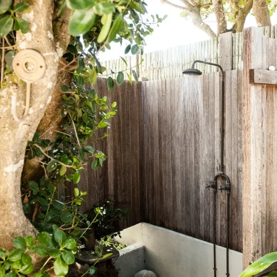 Outdoor shower enclosed by wooden cladding and positioned under a tree. Pebbles.