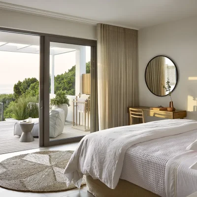 Walkers Beach Villa bedroom suite with a white waffle linen bed, a wicker floor rug, a small wooden desk and circular mirror, and glass sliding doors that open onto a wooden deck with a soft lounger and views of the swimming pool and ocean.