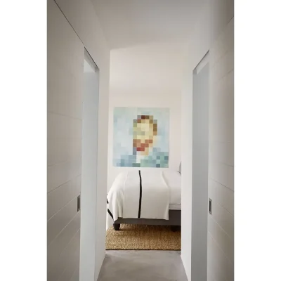 Bedroom passage with whitewashed walls that mimic the crisp, white linen on the bed and warmed by a woven floor rug with a pixelated avant- garde art piece. The Herenberg.