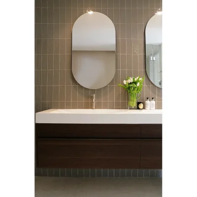 Tiled bathroom wall with two lit up, mounted oval mirrors, a large wooden cabinet with white ceramic sink and a vase of white tulips.