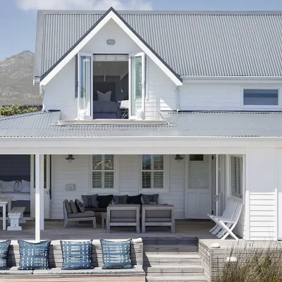 White beach house with large windows and glass doors, an expansive wooden deck with outdoor furniture and a backdrop of mountains with a clear blue sky. Noordhoek Beach House.