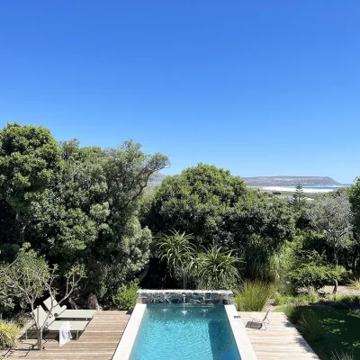 Back garden of Pinebeach with a large swimming pool surrounded by a wooden deck with sun loungers and surrounding luscious trees, and a view of Noordhoek beach in the distance.