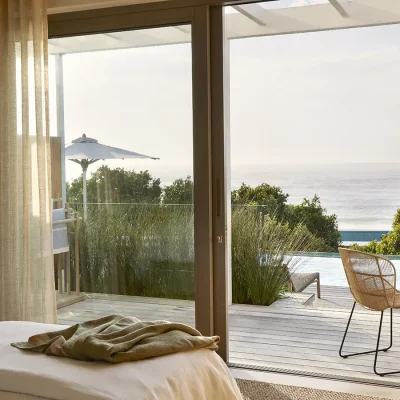 Large glass sliding doors with beige linen curtains revealing the wooden deck set with a small dining table and chairs, and overlooking the swimming pool and rolling ocean. Walkers Beach Villa.