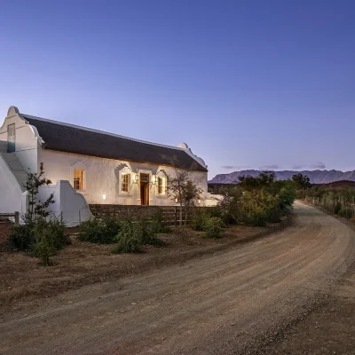 Buffelsdrift original main house with whitewashed walls, a thatched roof and lit up stoep situated next to a gravel road with a backdrop of the evening sky and mountains in the distance.