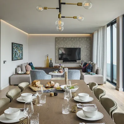 Open plan living and dining area with lengthy wooden dining table facing the lounge with beige walls that match the corner sofa's and two blue checkered armchairs facing a flatscreen television mounted on a beige decorative wall. Labotessa Hotel.