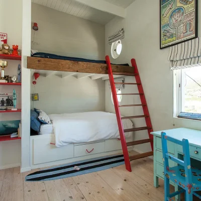 Child friendly room with a built- in bunk bed, white cladded walls, and touches of red in the ladder and floating book shelves that compliment the blue desk and blue pillows on the white bedding.