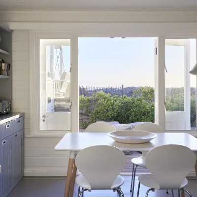 Noordhoek Beach house dining nook in the kitchen with a charcoal grey cabinet and a simple white table and chairs facing views of the garden and distant mountains.