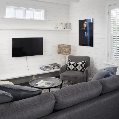 Secluded lounge with a soft charcoal 'L-shape' sofa and matching armchair facing a flat screen television mounted on white cladded walls with surrounding windows. Noordhoek Beach House.