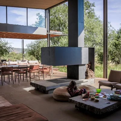 Spacious open planing living area with an open fireplace that is central to the large wooden dining table and leather armchairs, and surrounded by glass stack back doors that reveal the natural landscape of Witklipfontein eco lodge.