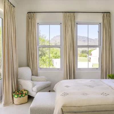 Comfortable bedroom with beige hues, a spacious bed with white linen and beige throw, a striped corner armchair and surrounding windows and double doors with views of Buchubos villa garden.