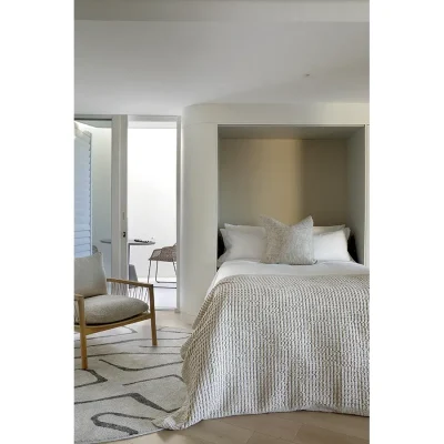 Main suite in Ebb Tide with a luxurious 'murphy bed', a textured cream floor rug and a comfortable, cream arm chair that is located in front of large glass sliding doors that open onto a private balcony.