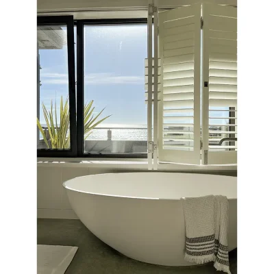 Bathroom with white oval bath tub positioned adjacent to large stack back glass windows with white shutters exposing views of the ocean. Oasis On Glen.